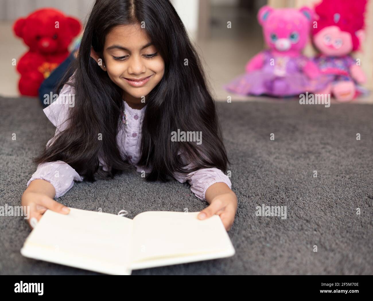 Portrait of a cute Indian school girl relaxing on a play mat, smiling and reading a diary book surrounded by toys. Stock Photo
