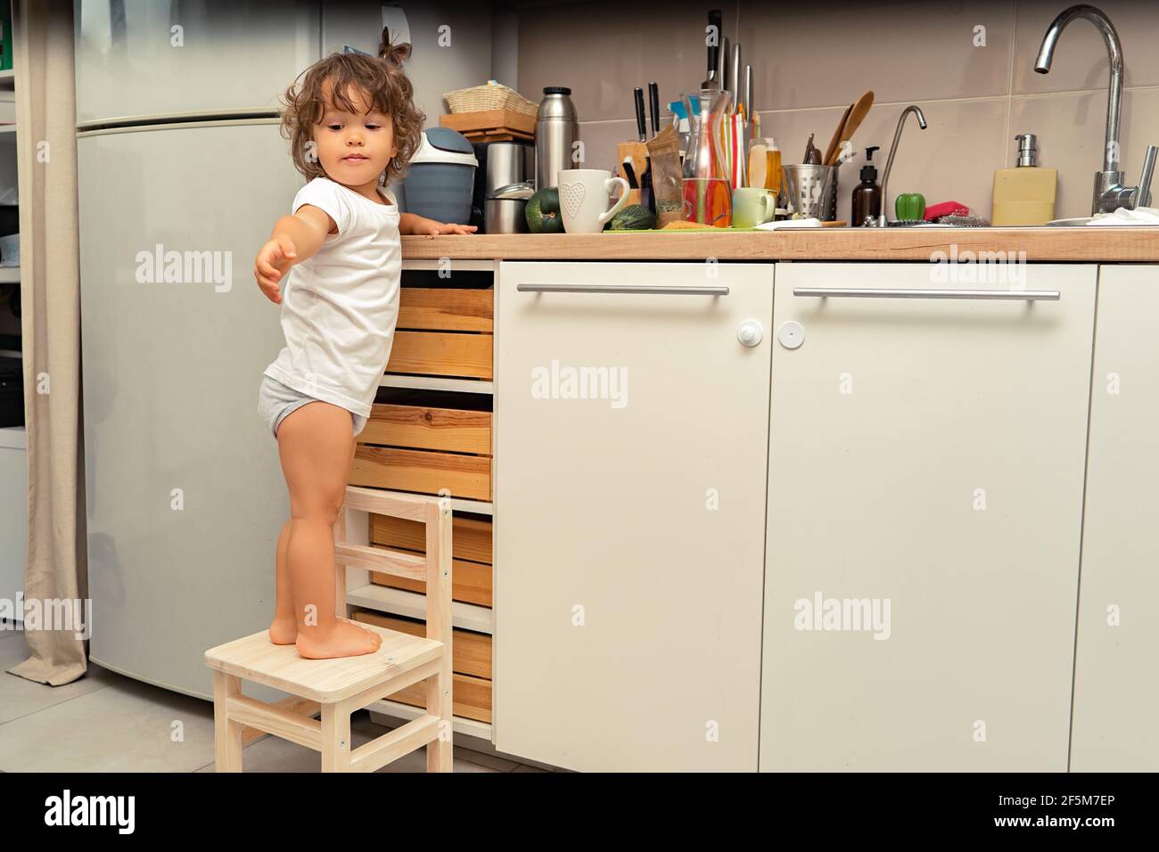 Little boy with long hair standing on a chair in the kitchen and asks for help Stock Photo