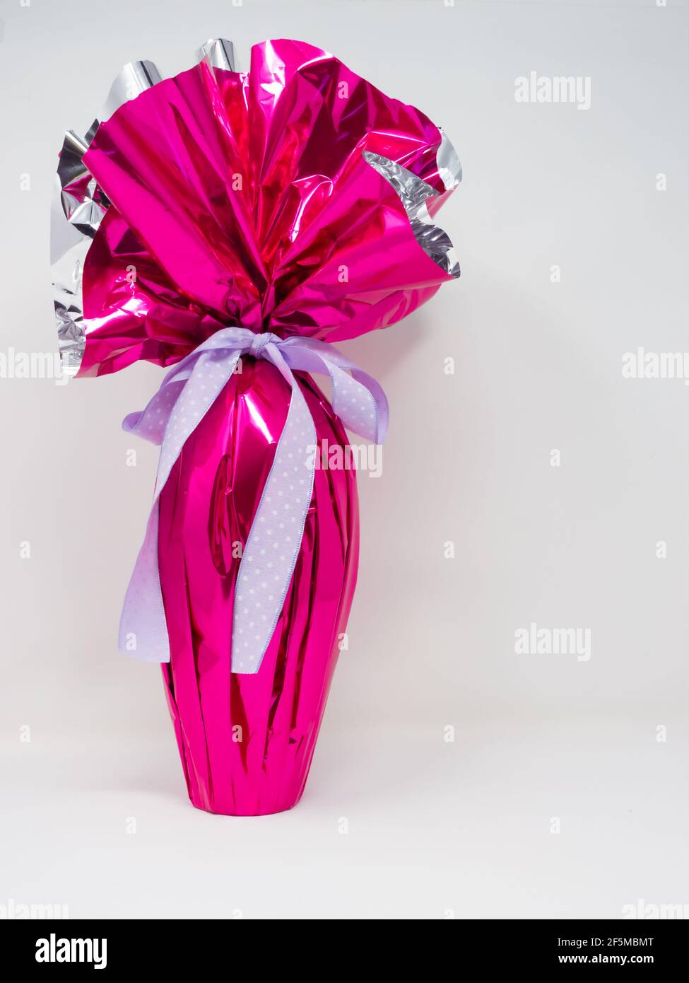 Giant chocolate Easter egg wrapped in pink silver foil with a pretty purple polka dot bow. Vertical, white background Stock Photo
