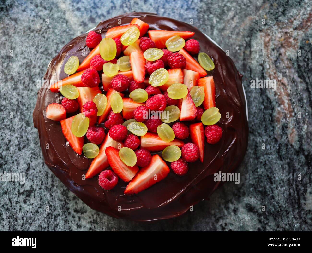 Top-Down Dark Chocolate Cake with Fruit on Marble Kitchen Counter. Tart with Chocolate Icing and Strawberries, Grapes and Raspberries. Stock Photo