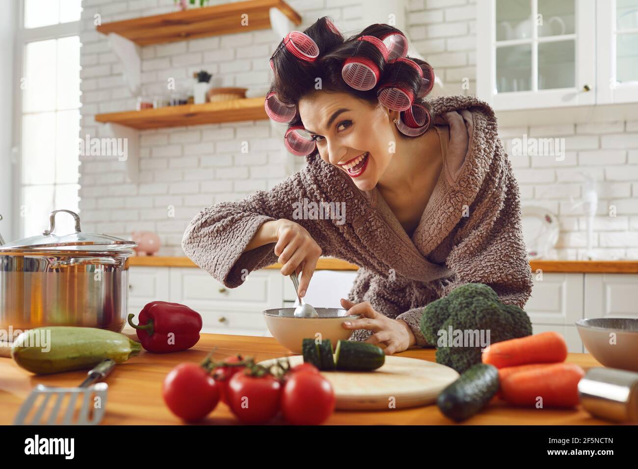 Funny woman housewife in curlers and bathrobe standing cooking fresh healthy dish Stock Photo