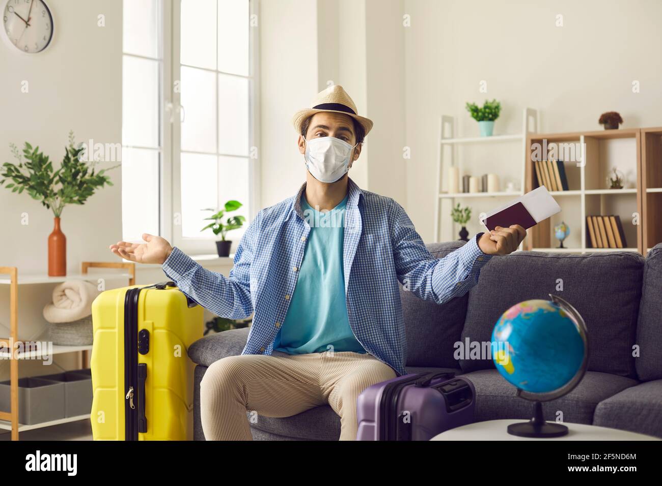 Upset man in face mask sitting with luggage and flight tickets at home Stock Photo