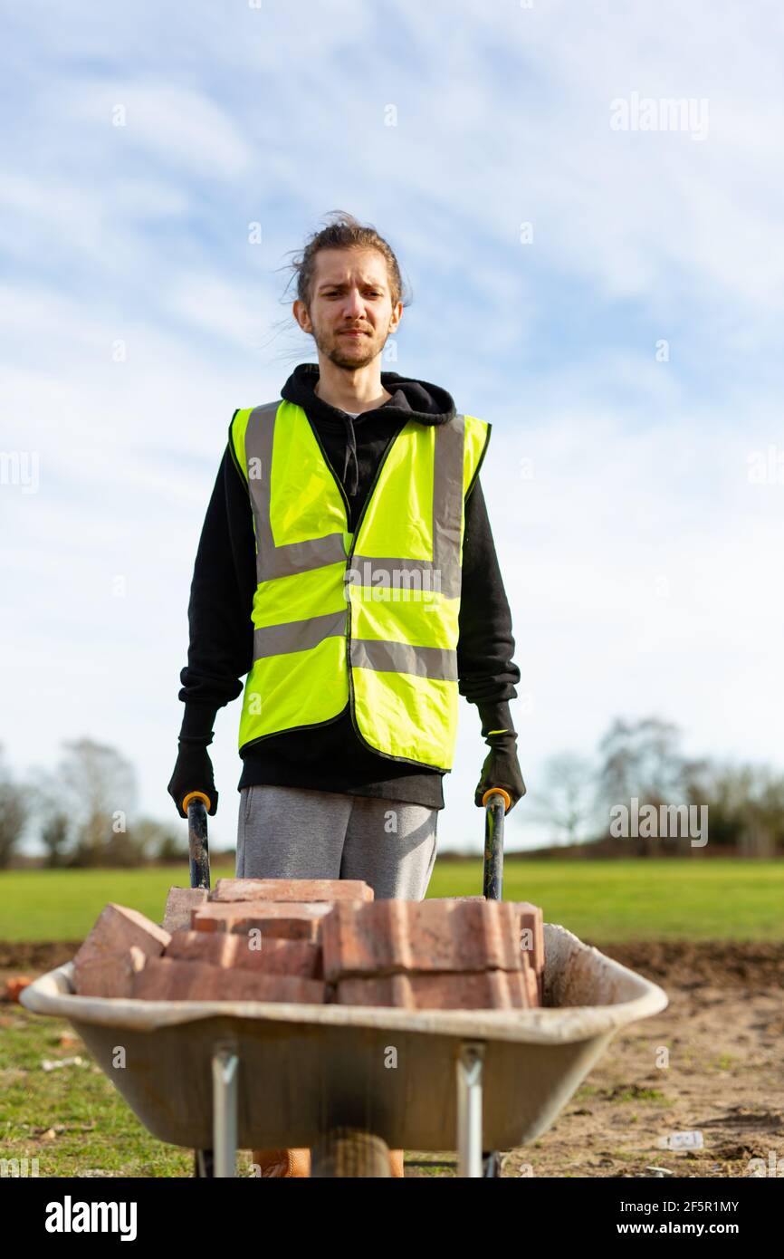 A young adult male builder wearing a high visibility vest and hard hat pushing a wheelbarrow full of bricks while on a building site Stock Photo