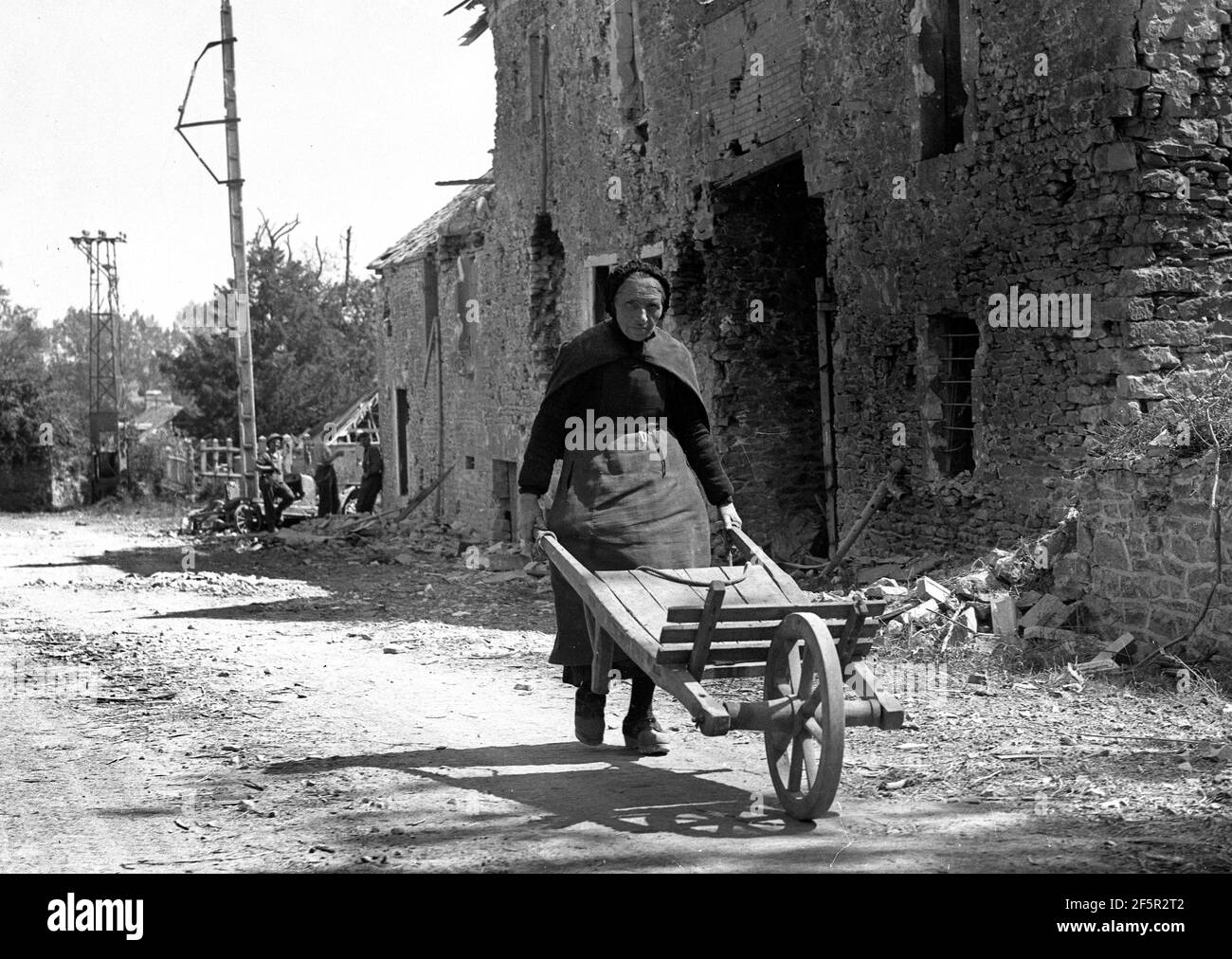 Rural France World War Two 1944. Old lady pushing hand cart through French village past war damaged buildings with allied soldiers in the background during the liberation of France Stock Photo