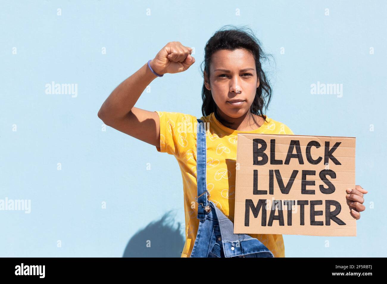 Portrait of Latin American woman with serious attitude and raised fist. She is isolated on blue background holding a banner with slogan Black Lives Ma Stock Photo