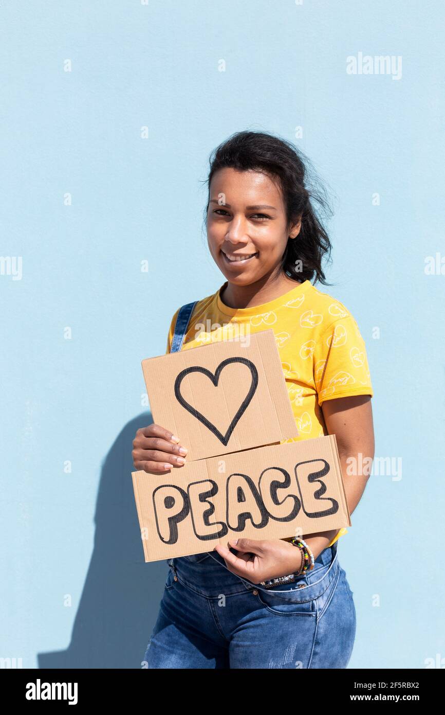 Portrait of beautiful hispanic woman with tender gesture holding a banner with text Peace. She is isolated on blue background. Space for text. Stock Photo