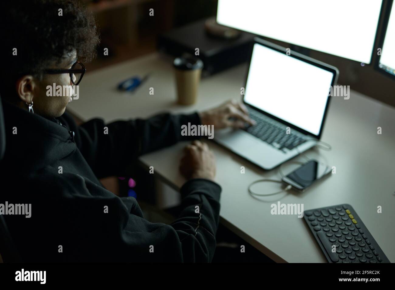 Young guy wearing glasses sitting at the table in front of many computer monitors and using laptop while working late at night Stock Photo