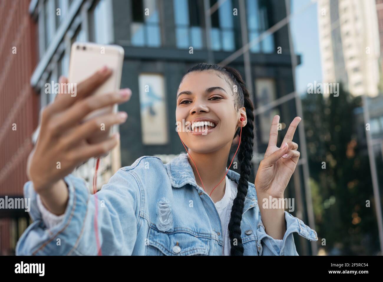 Beautiful African American woman with stylish hairstyle taking selfie, showing victory sign. Influencer using mobile phone streaming video Stock Photo