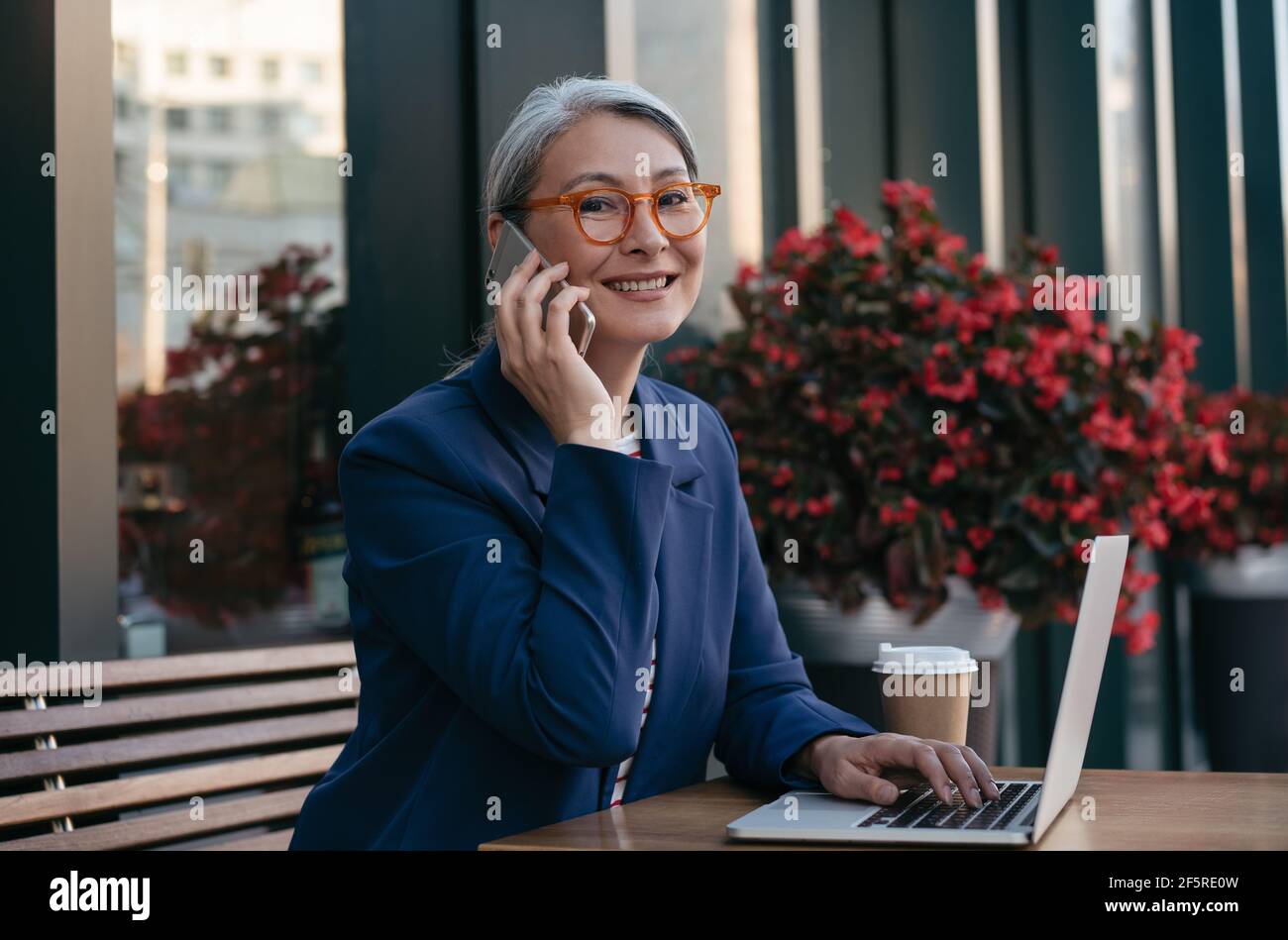 Mature asian woman wearing stylish eyeglasses talking on mobile phone, sitting at workplace. Portrait of middle aged businesswoman using laptop Stock Photo