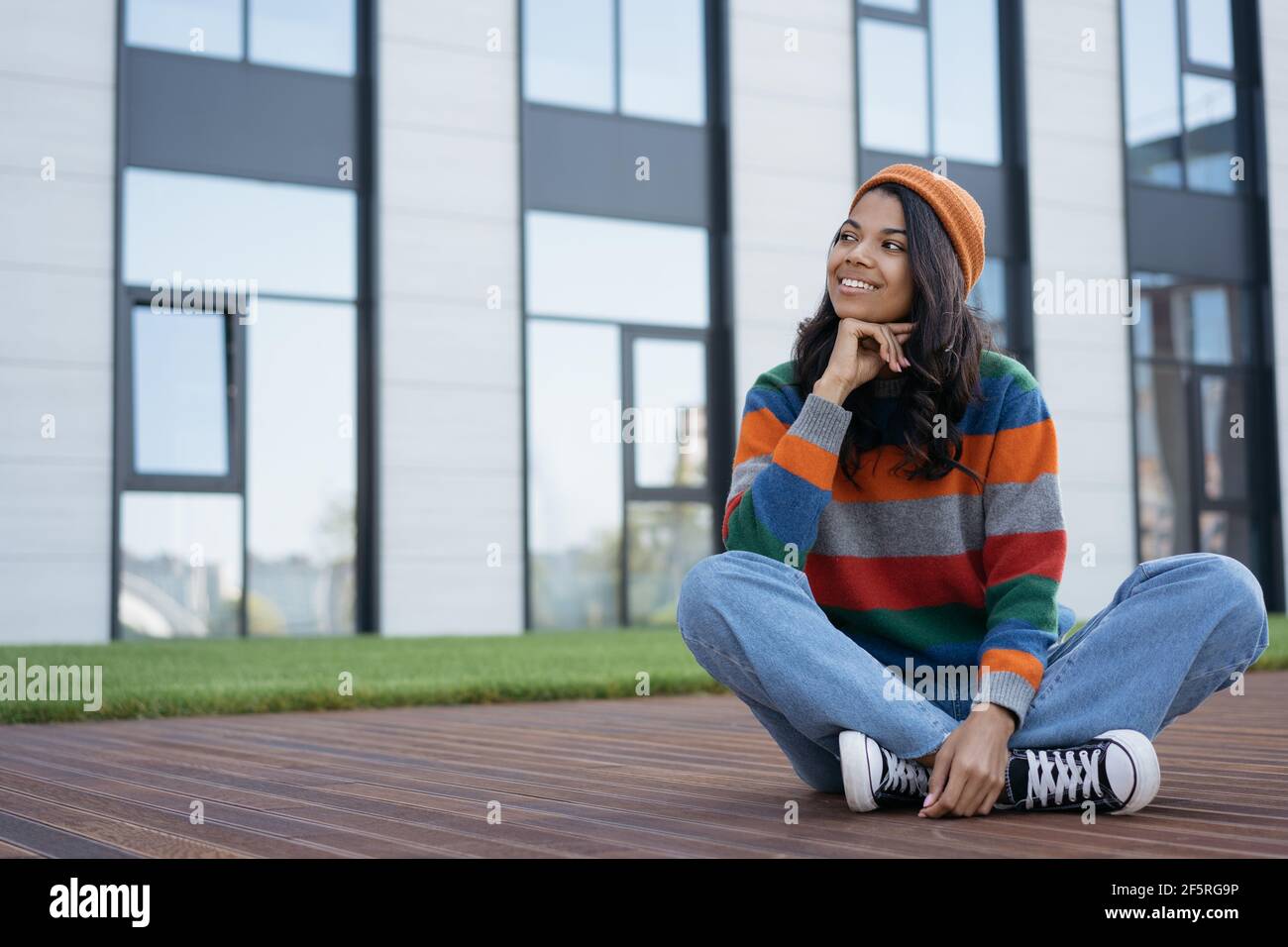 Beautiful smiling African American woman relaxing outdoors. Portrait of happy hipster wearing casual clothing and orange hat sitting in park Stock Photo