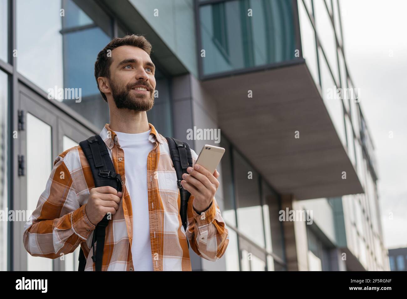 Smiling handsome man using mobile phone outdoors. Young bearded tourist with backpack walking on urban street, searching best way. Travel concept Stock Photo