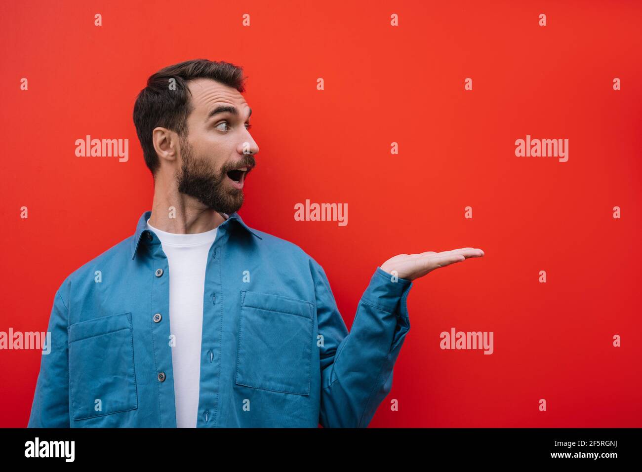 Emotional bearded man wearing casual shirt pointing with hand, isolated on red background. Copy space, advertising concept Stock Photo