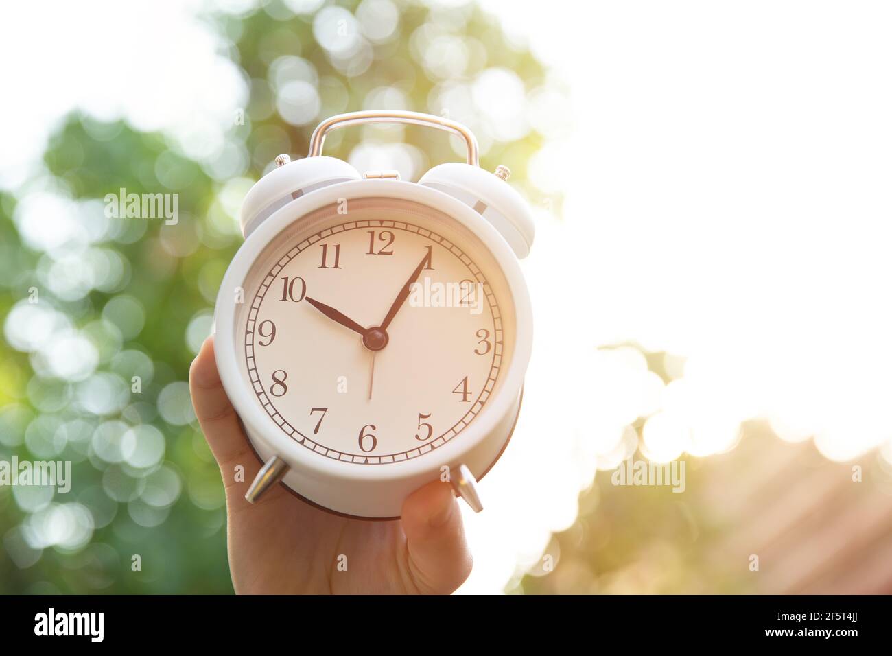 Morning time concept, hand hold white bell clock against nature sun