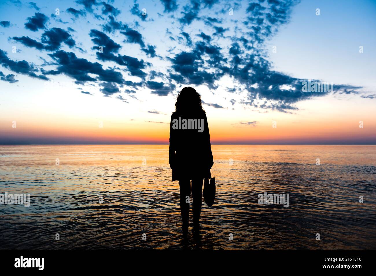 Back view of silhouette of unrecognizable female standing in water of sea and admiring spectacular view of colorful sundown sky in summer Stock Photo