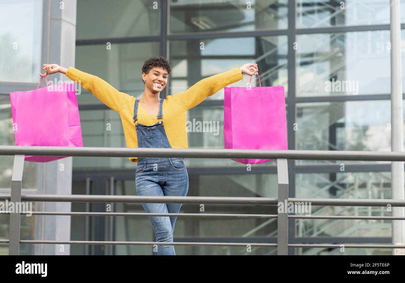 Delighted African American female standing with paper bags in city and looking at camera after successful shopping Stock Photo