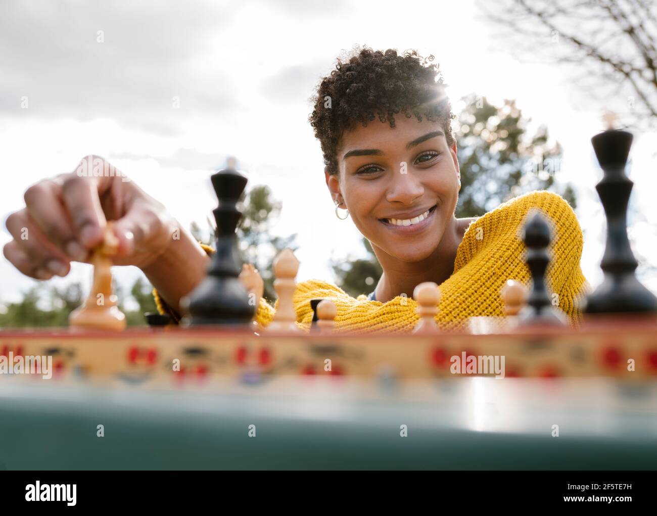 From below of optimistic African American female sitting at table in park and playing chess while looking at camera Stock Photo