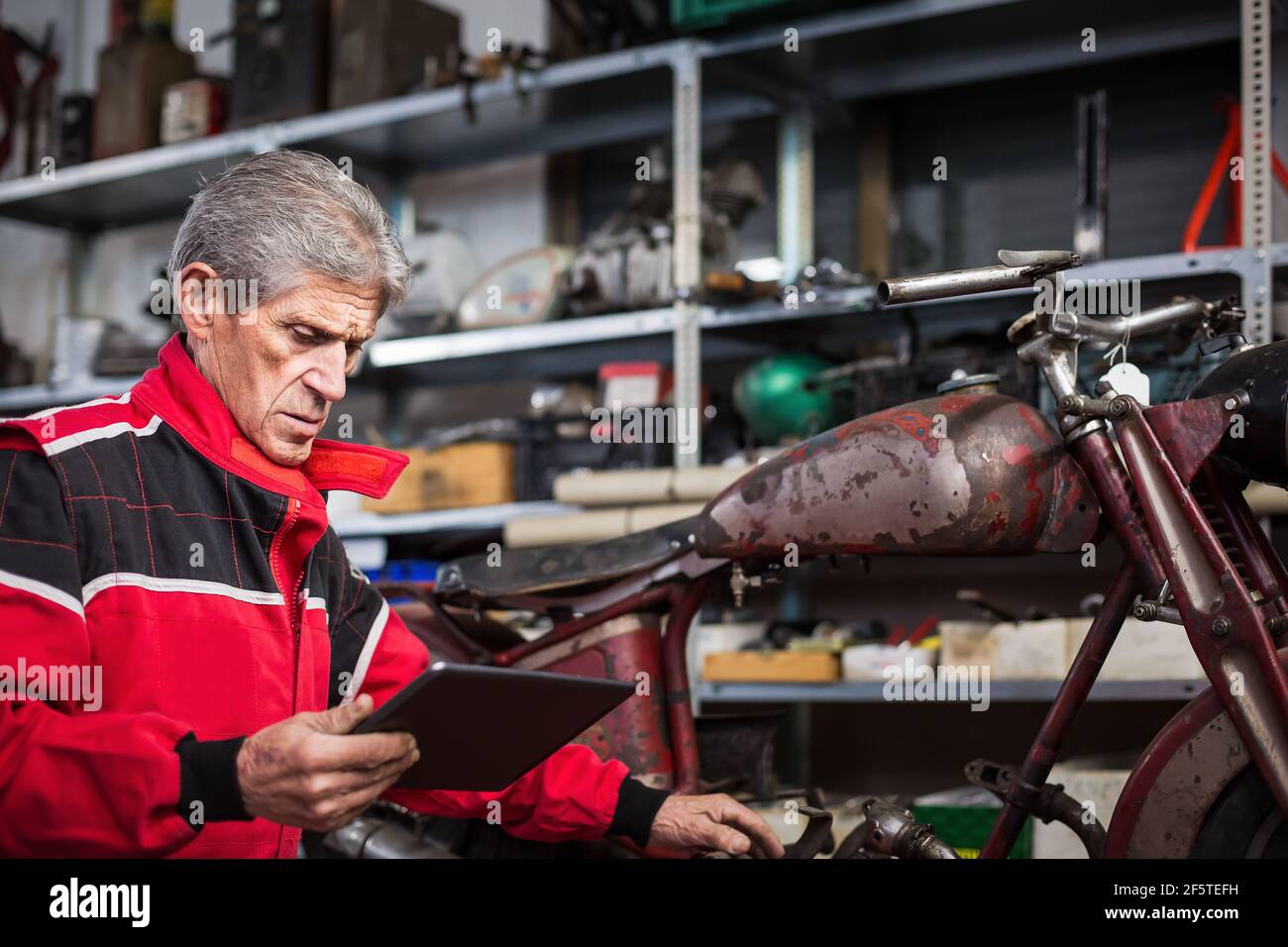 Serious gray haired elderly repairman using tablet and watching video instruction while fixing old broken motorcycle in garage Stock Photo