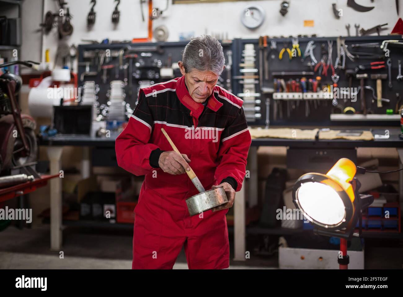 Skilled senior repairman in workwear mixing paint in metal pot during repair works in moto service workshop Stock Photo