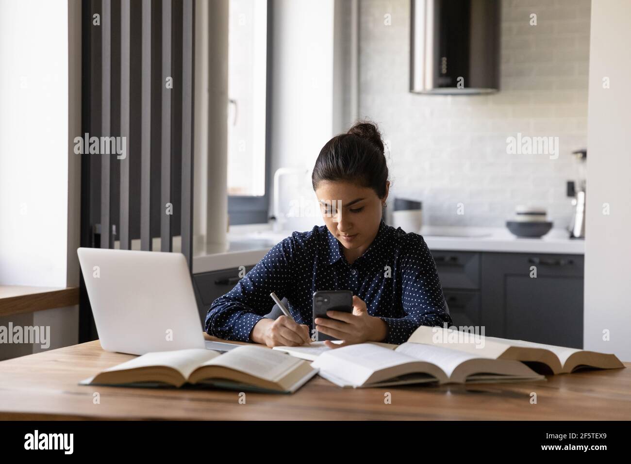 Focused Indian woman holding phone, writing notes, studying online Stock Photo