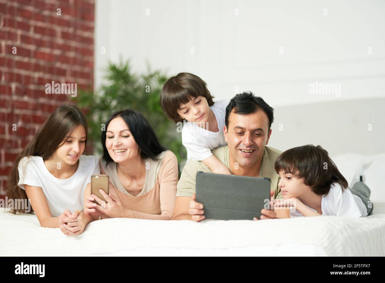 Boys and girls. Happy latin family with cute kids using digital devices, lying on the bed together Stock Photo