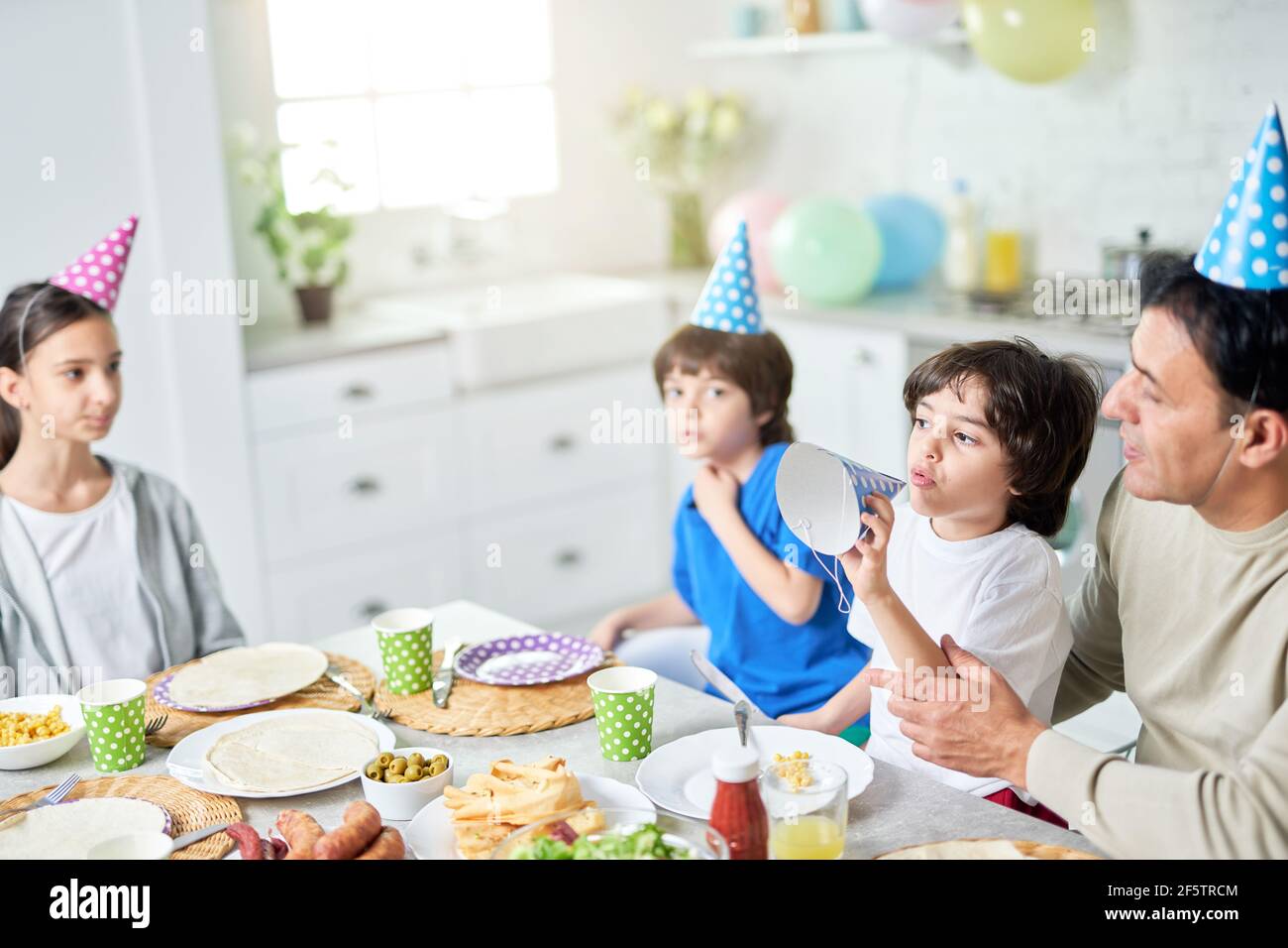 Carefree athmosphere. Joyful little boy playing with his birthday cap. Hispanic family having dinner while celebrating birthday together at home Stock Photo