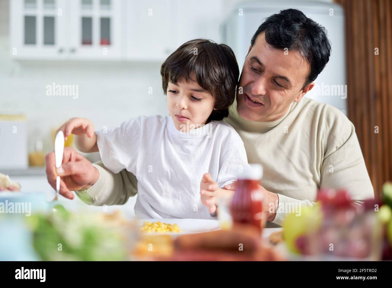Caring Dad. Smiling middle aged latin father serving his son, sitting with him at kitchen table while having a lunch at home Stock Photo