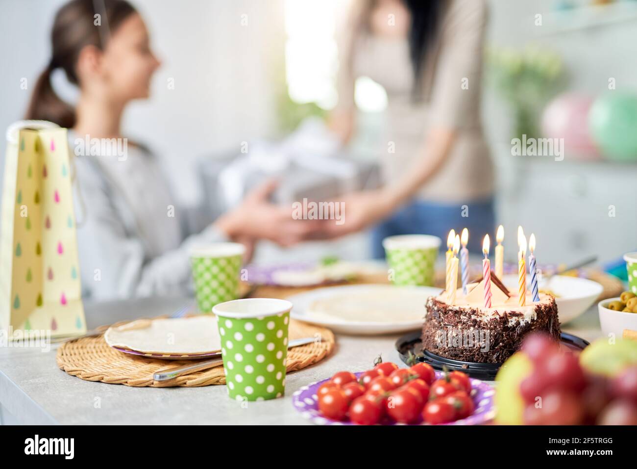 Greetings. Close up of a birtday cake on the table. Mother giving present to her daughter, celebrating birthday in the background Stock Photo