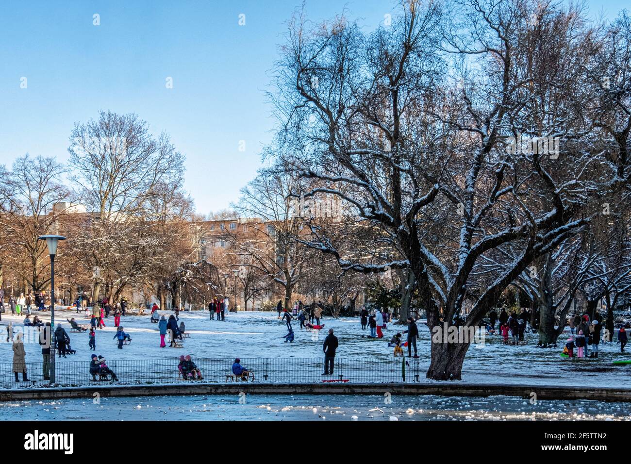 Parents watch children tobogganing next to a frozen pond in snow covered Weinbergs Park, Mitte, Berlin Stock Photo