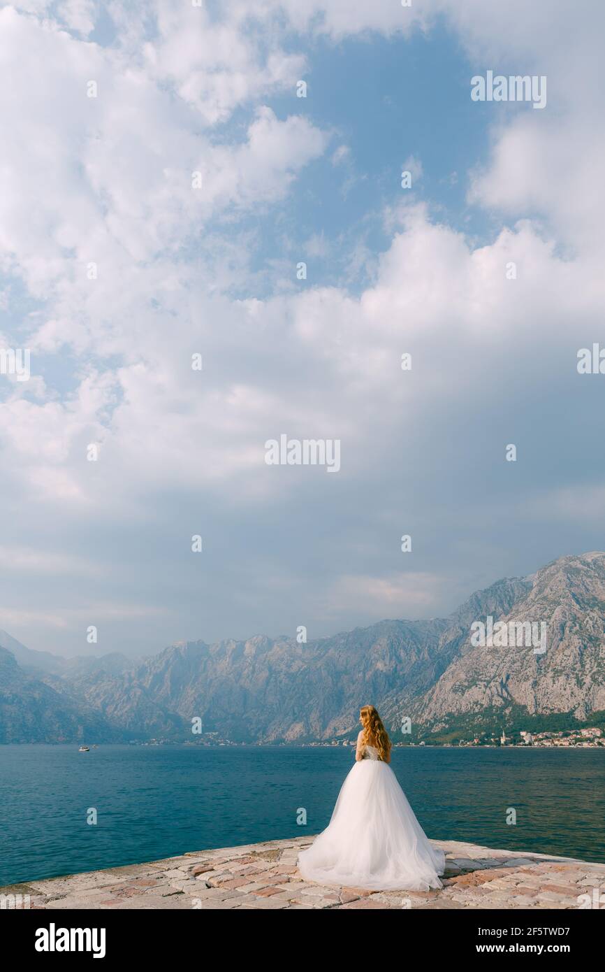 A beautiful blonde bride in a puffy white dress stands on a pier in the ...