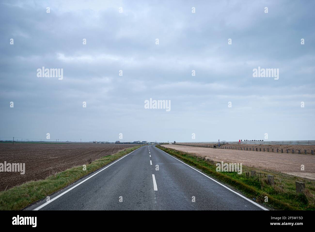 single point perspective of two lane black top road stretching to horizon through flat farmland with buildings faintly in the disctance. Stock Photo