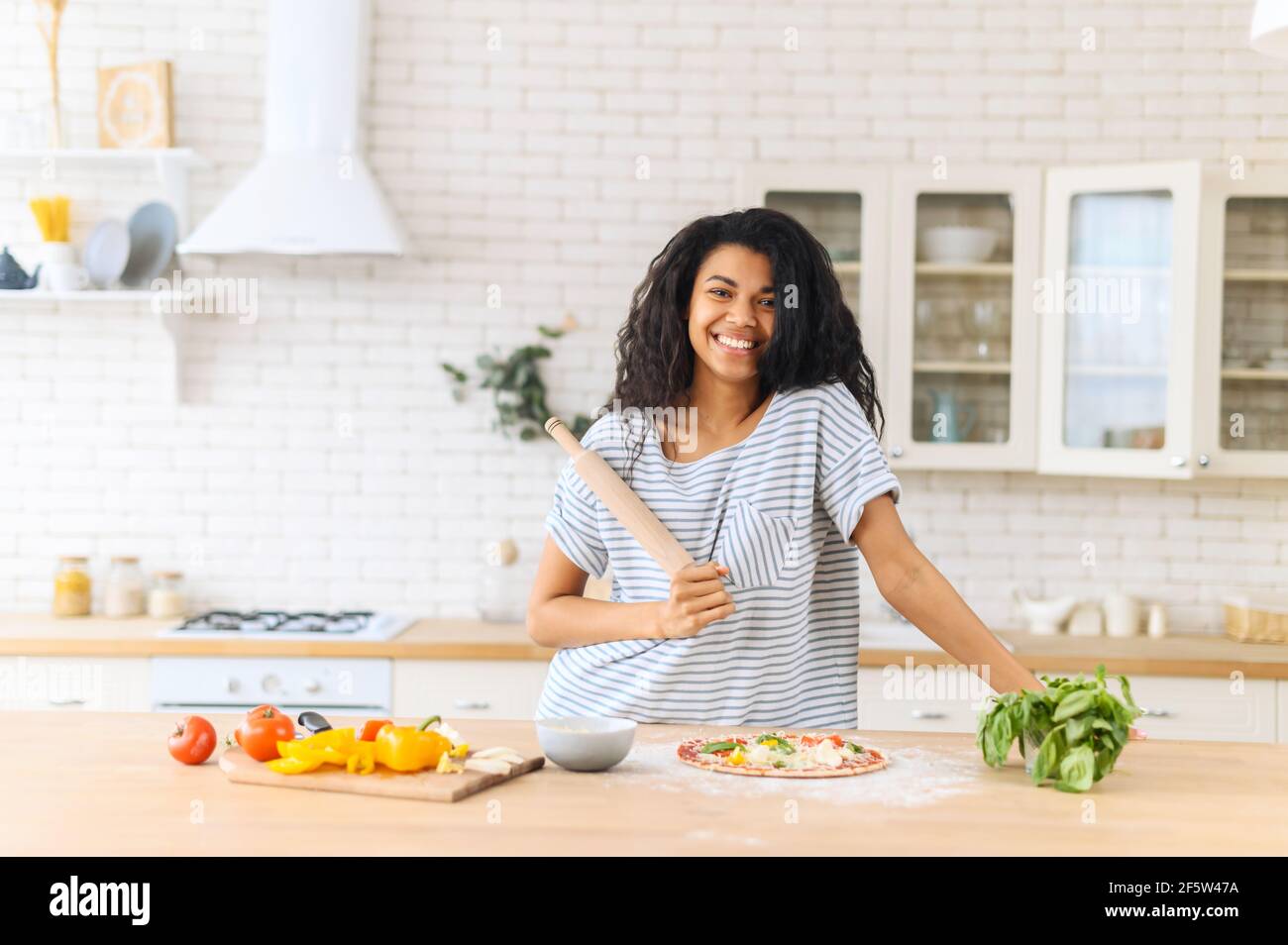 Happy attractive biracial adult woman housewife wear wide T-shirt holding rolling pin on kitchen table baking pizza concept, cooking healthy homemade pizza with a lot of greens and vegetables at home Stock Photo
