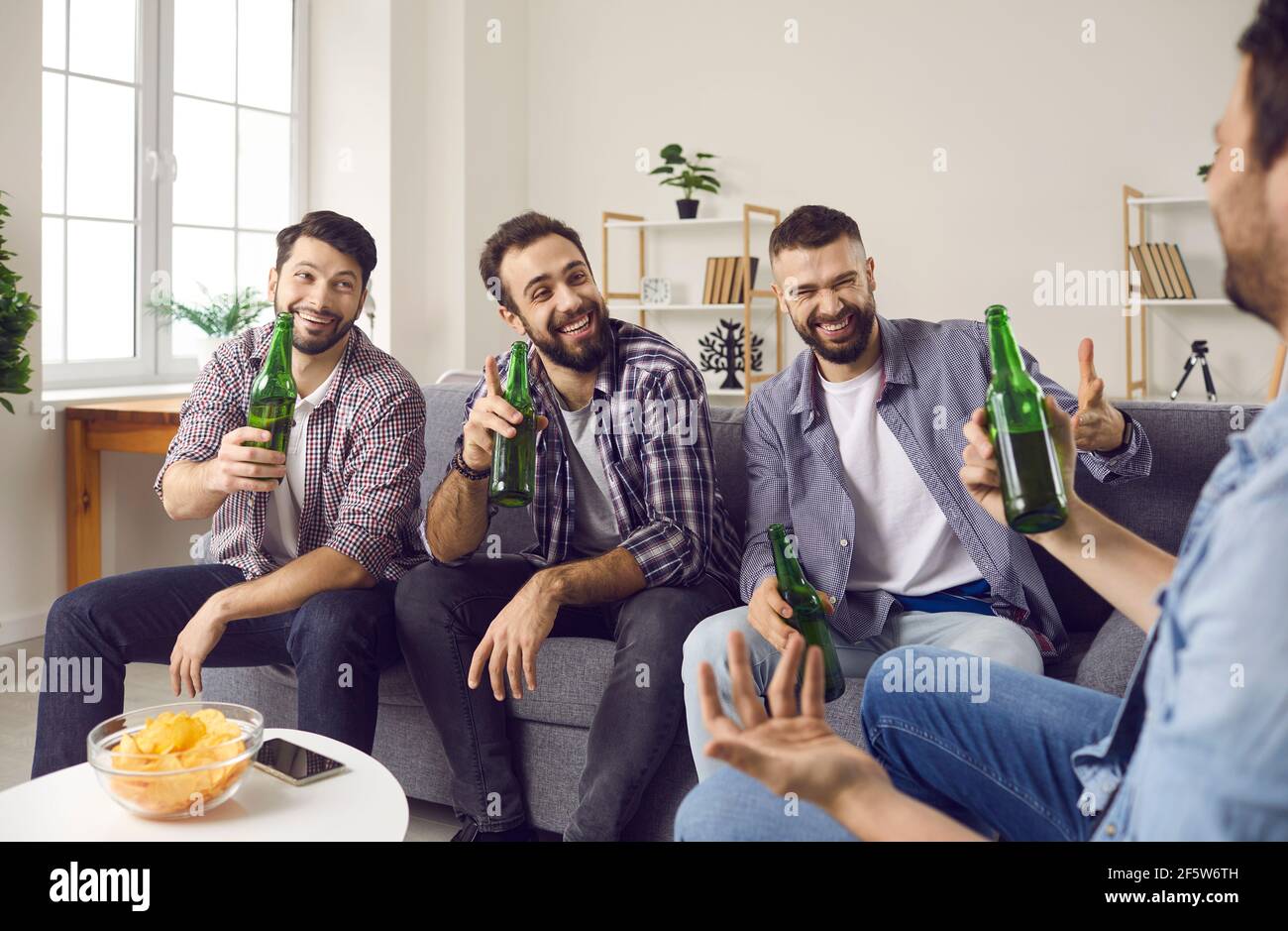 Group of happy young friends sitting on couch, drinking beer and telling funny stories Stock Photo
