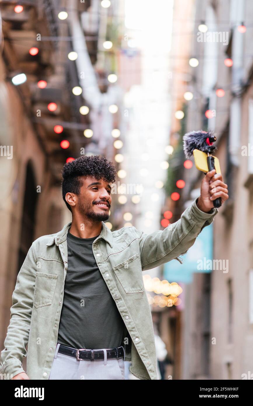 latin american young man holding a selfie stick with phone and microphone and recording himself. He is walking on the streets and smiling to phone Stock Photo