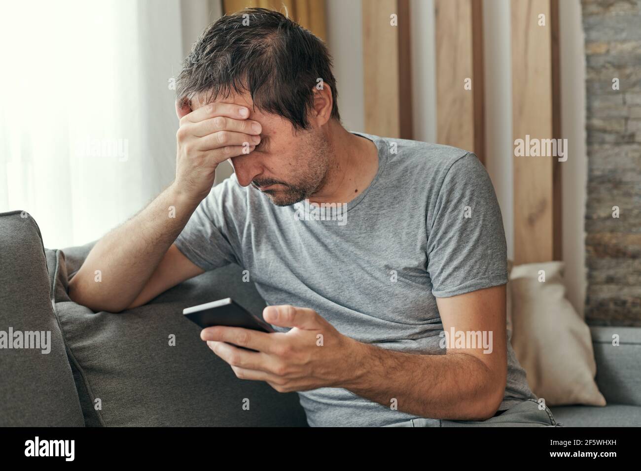Troubled man receiving bed news in text message on mobile phone while sitting at living room sofa Stock Photo