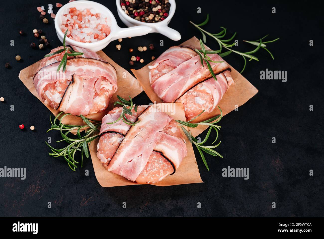 Three chicken hamburgers wrapped in bacon on cave paper with rosemary branches, black pepper and pink salt in white small plates, black background. Fa Stock Photo