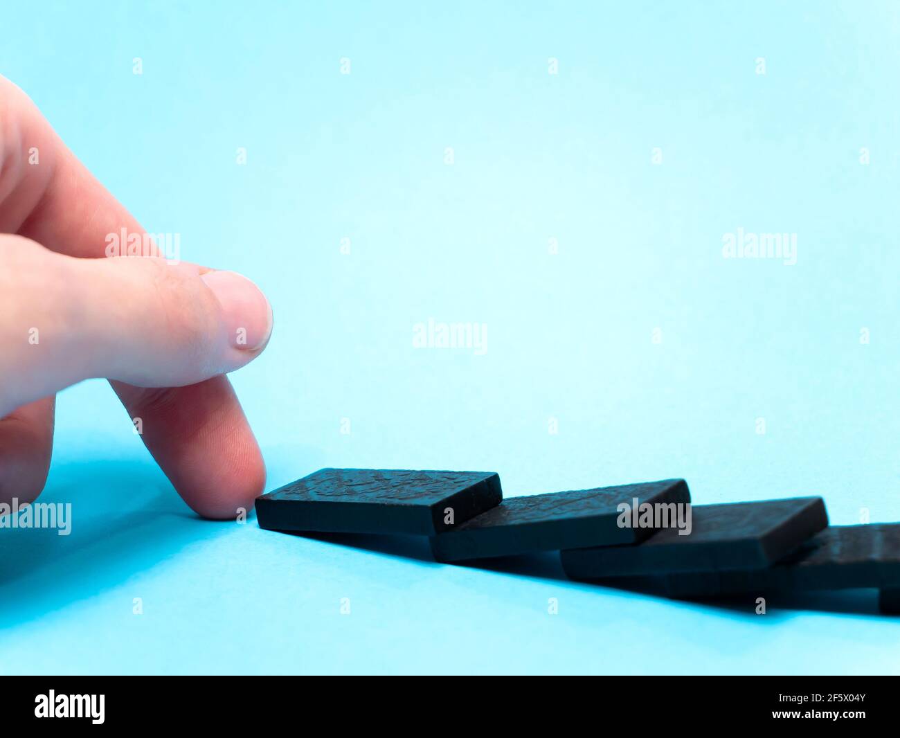 Domino effect, hand closeup. Finger knocked over a row of domino pieces, dominoes fallen over, laying on a blue background. Knocking over, toppling do Stock Photo