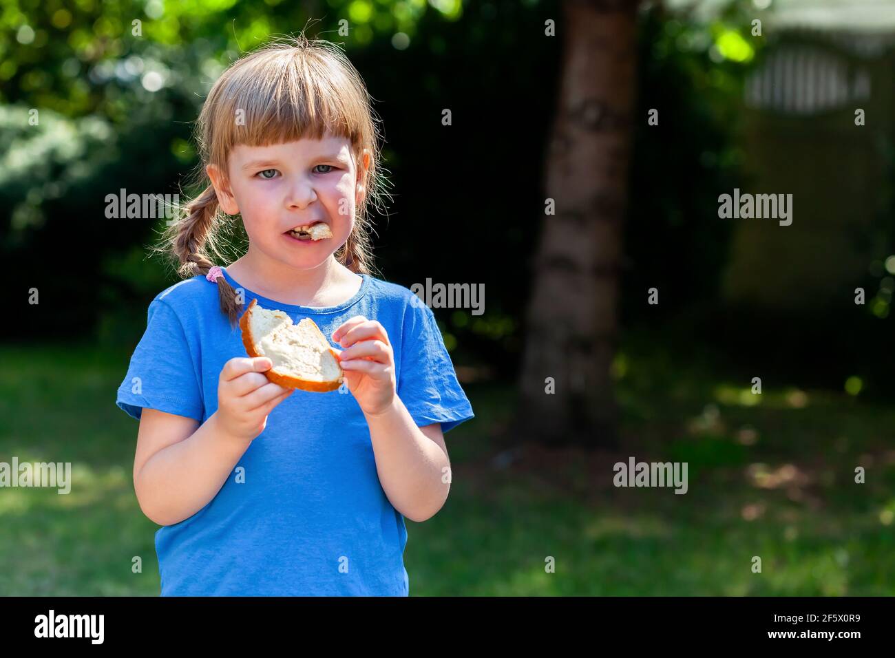 One little school age girl eating a single slice of bread with butter outdoors, copy space. Young kid taking a bite of a bread slice alone, nutrition, Stock Photo