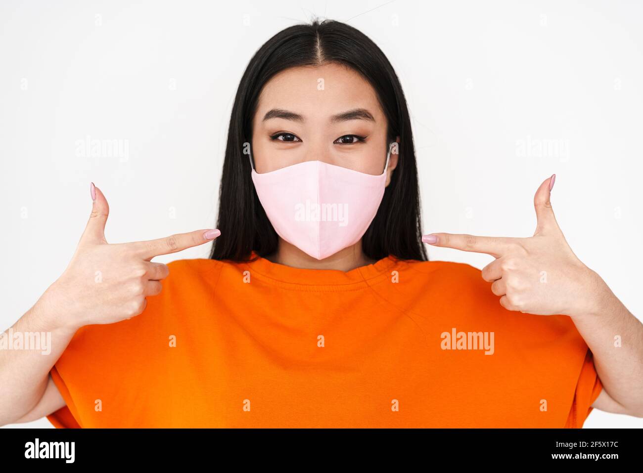 Asian Pleased Girl In Face Mask Pointing Fingers At Her Face Isolated asian-pleased-girl-in-face-mask-pointing-fingers-at-her-face-isolated