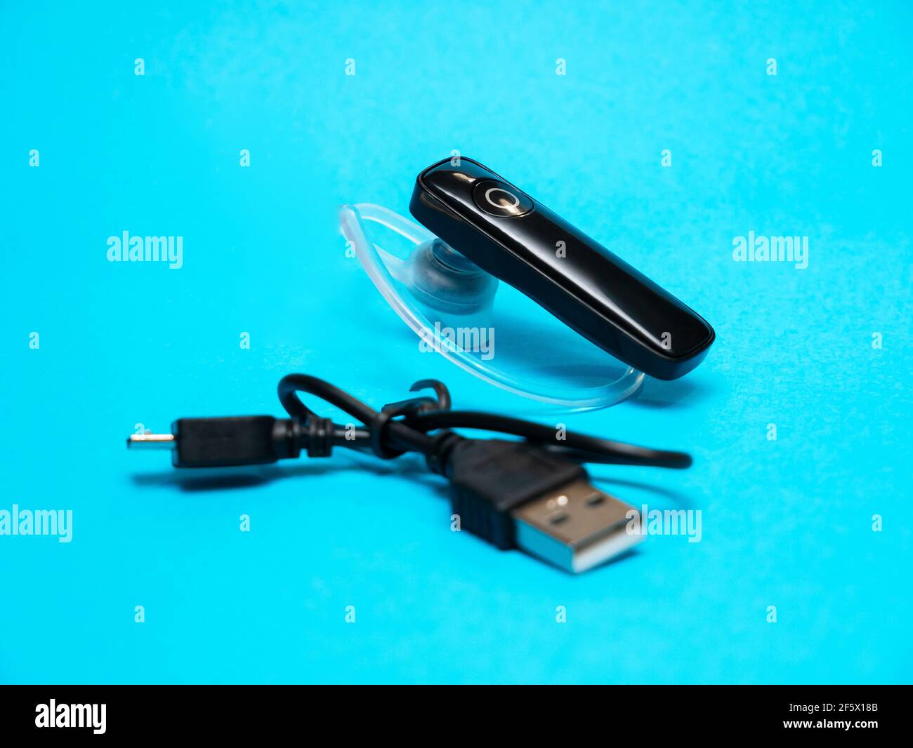 Simple hands-free wireless earpiece with a usb charging cable, object on a blue background, closeup, studio shot. Ergonomic mono handsfree headset wit Stock Photo