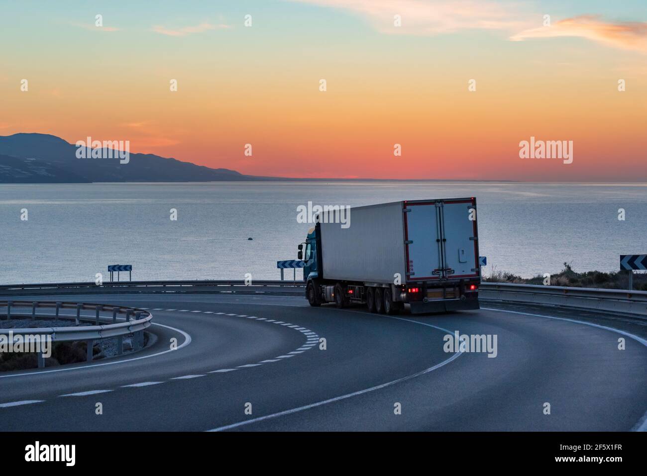 Truck with refrigerated semi-trailer on a mountain road with the sea and the sun on the horizon. Stock Photo