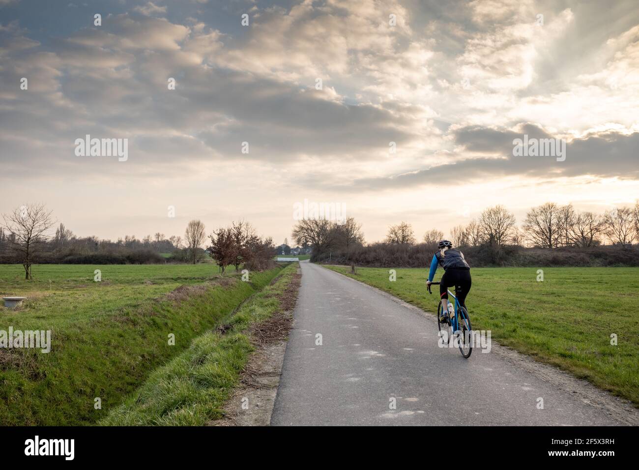 Outdoor sunny view of cyclist ride a bicycle on small road in suburb area surrounded with agricultural field in spring season against dramatic sky. Stock Photo