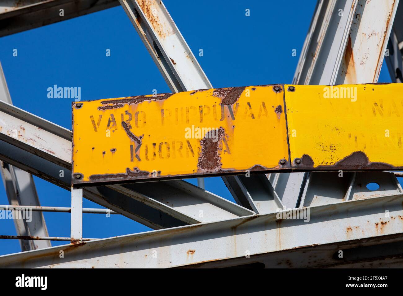 Old yellow rusty warning sign on dock crane structure against clear blue sky in Jätkäsaari or Länsisatama district in Helsinki, FInland Stock Photo