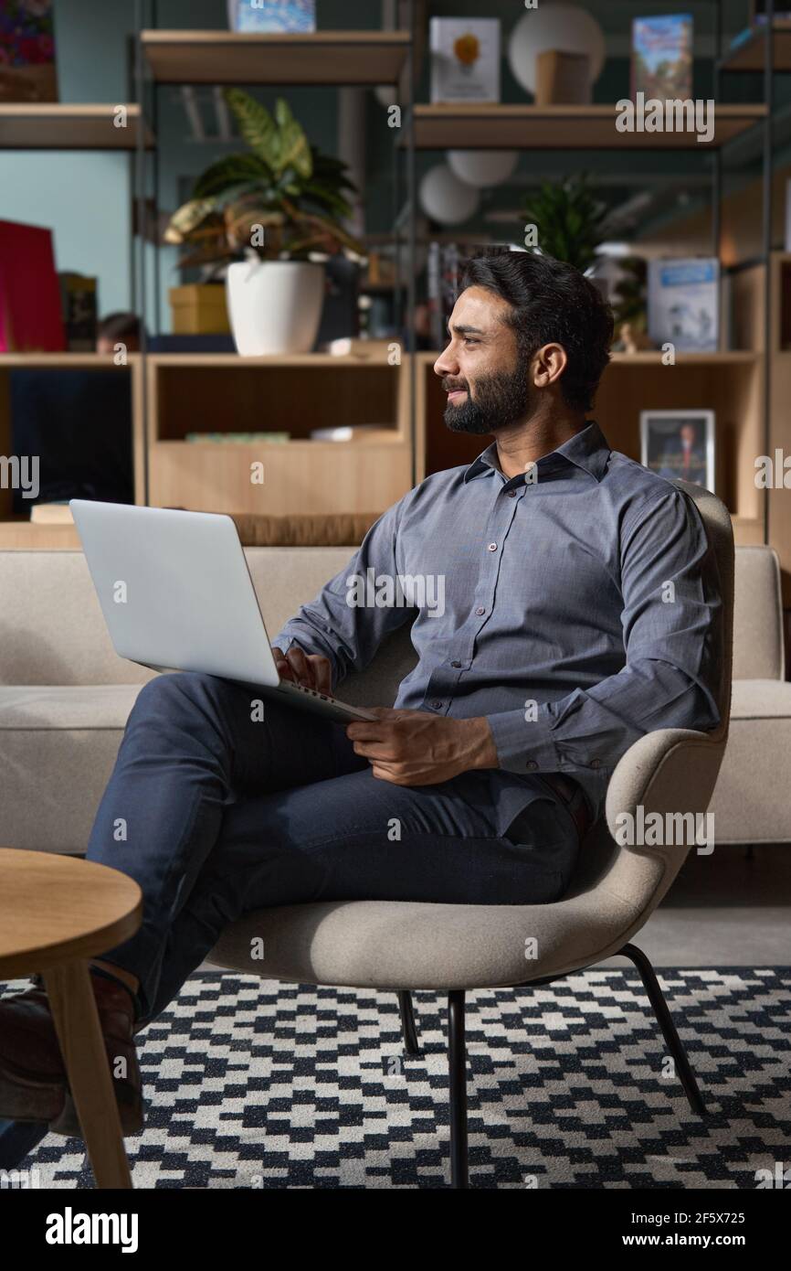 Indian business man looking away sitting on chair in modern office using laptop. Stock Photo