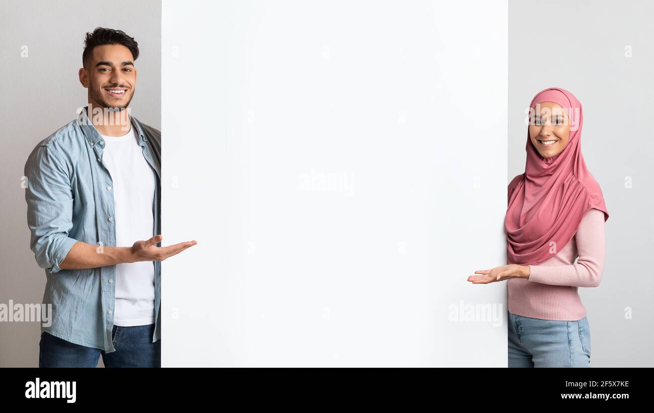 Nice Offer. Smiling muslim couple pointing at white advertisement board with hand Stock Photo