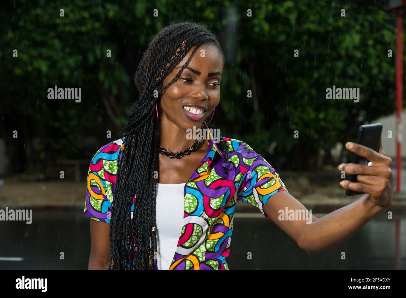 young african girl under a light rain watching mobile phone while ...