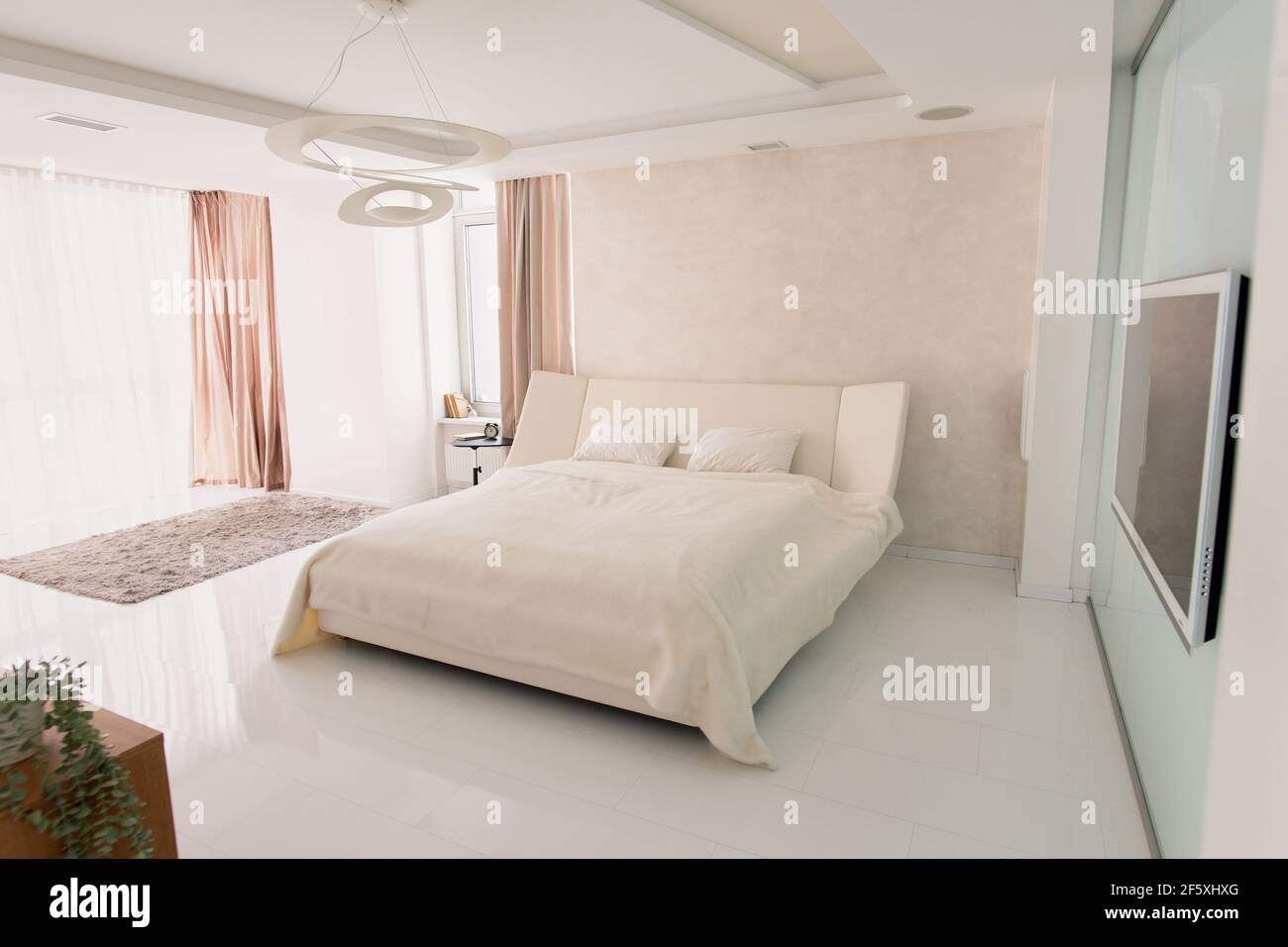 Interior of large cozy bedroom with comfortable double bed, plasma tv on wall, alarm clock on small table and grey furry rug on the floor Stock Photo