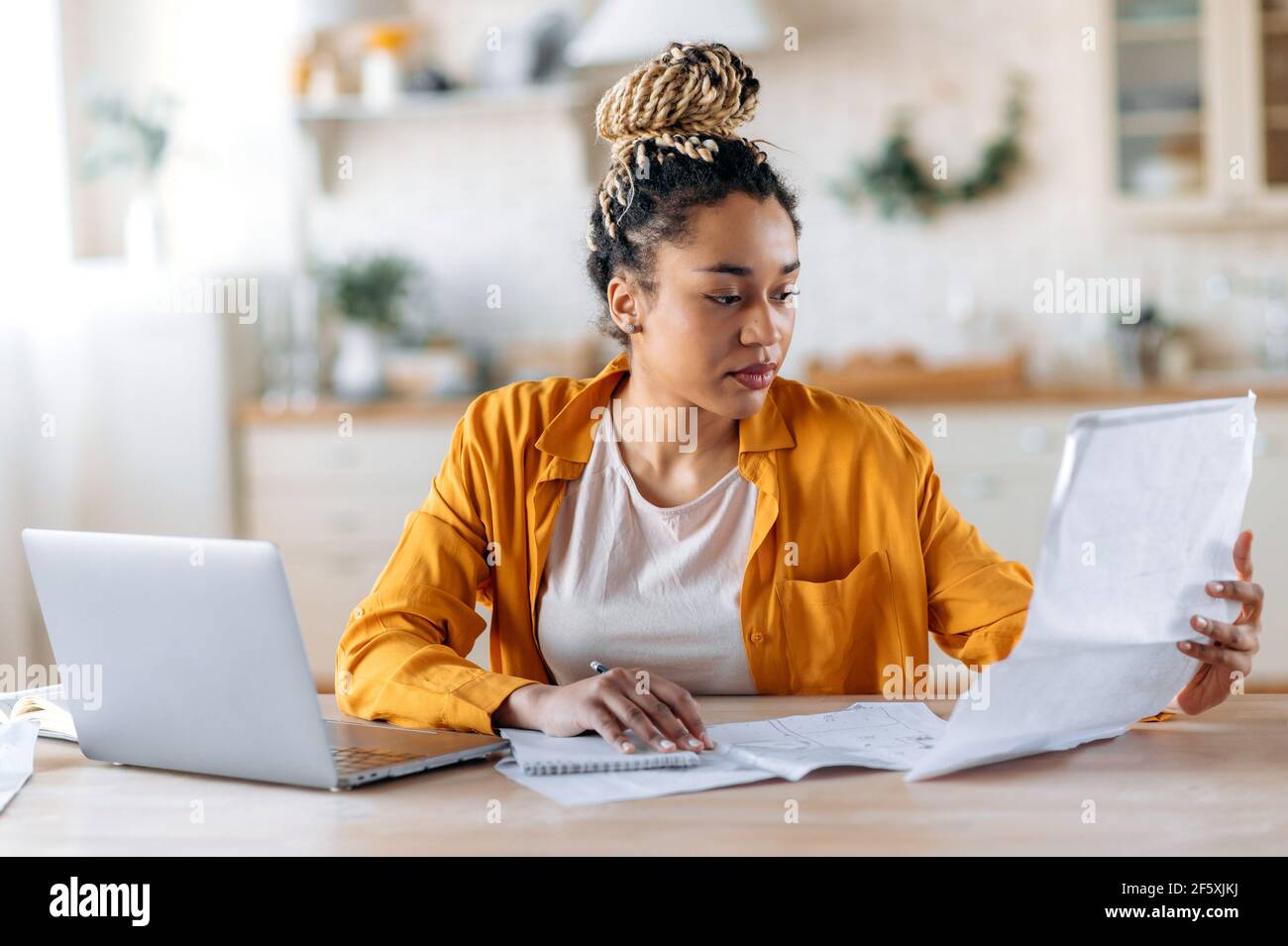 Concentrated clever african american woman, student or designer, wearing stylish casual clothes, studying or working remotely, using laptop, watching and examines blueprints. Work from home Stock Photo