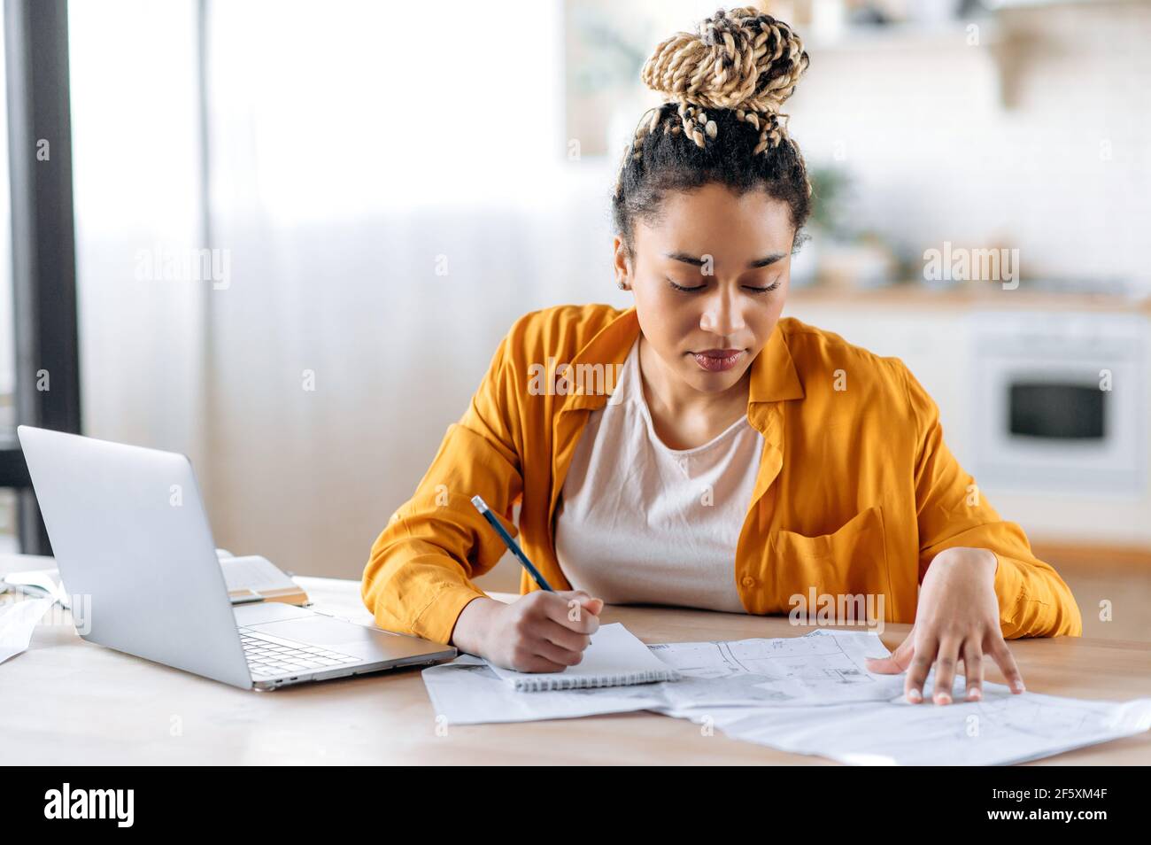 Confident smart focused african american female student or designer wearing stylish casual clothes, studying or working remotely, using laptop, taking notes, examines blueprints. Work from home Stock Photo