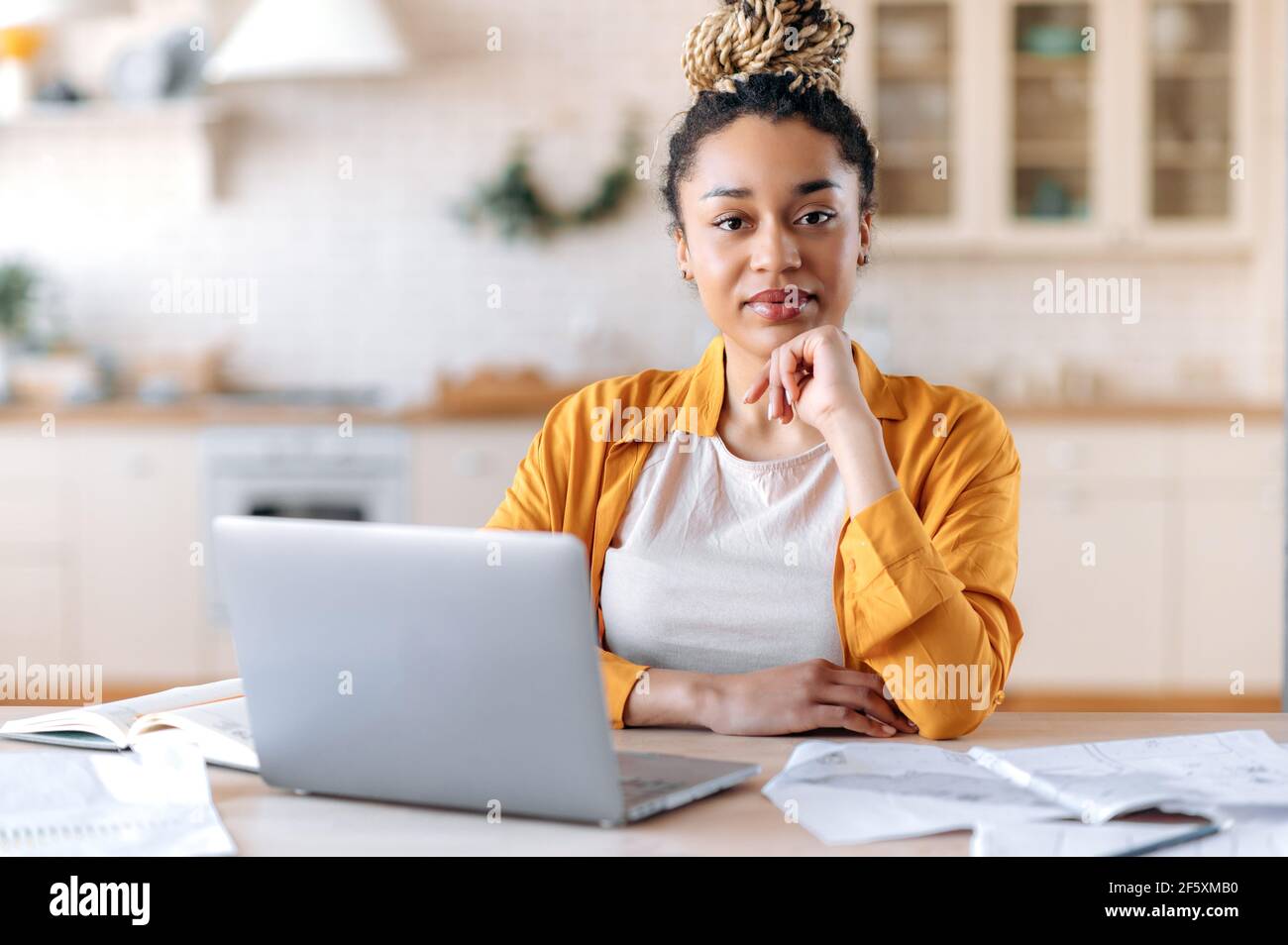 A serious intelligent young african american woman with dreadlocks, manager, freelancer or real estate agent in casual clothes, sits at the work desk, working from home, looks at camera Stock Photo