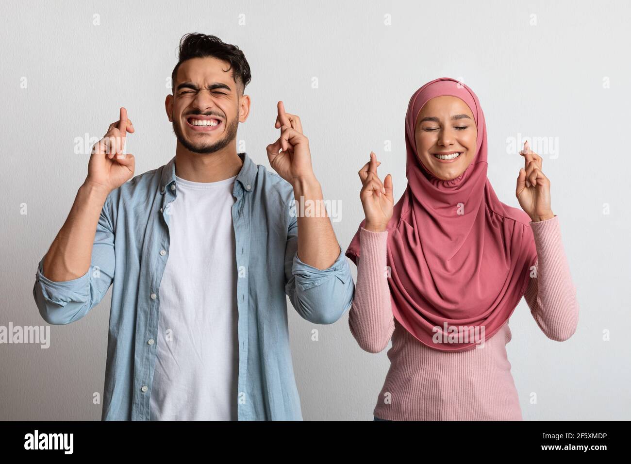 Making Wish. Superstitious muslim couple crossing fingers for luck with closed eyes Stock Photo